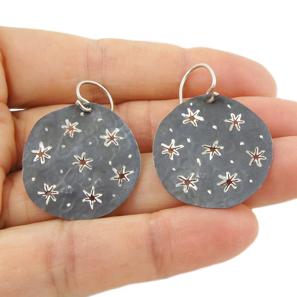 Round silver earrings with a black finish and star designs held between fingers on a white background