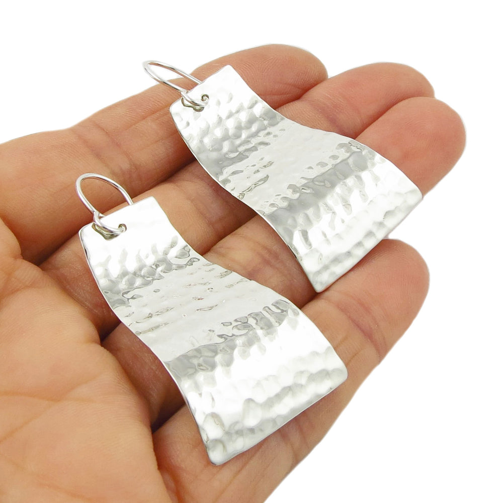 A pair of long curving 925 silver rectangle earrings with a hammered finish held in a person's hand on a white background.