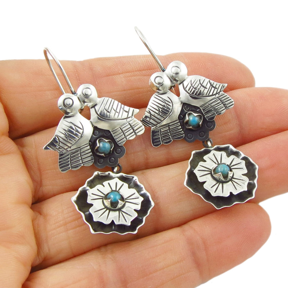 A pair of silver earrings featuring a shadow box design with doves and flowers, and turquoise gemstones at the center, held in a person's hand.