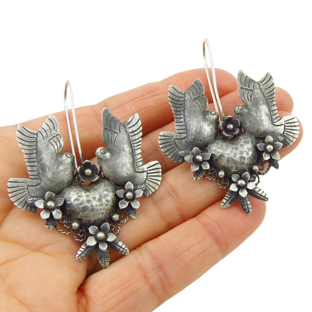 Blackened Taxvo silver lovebird and flower earrings held in a person's hand.