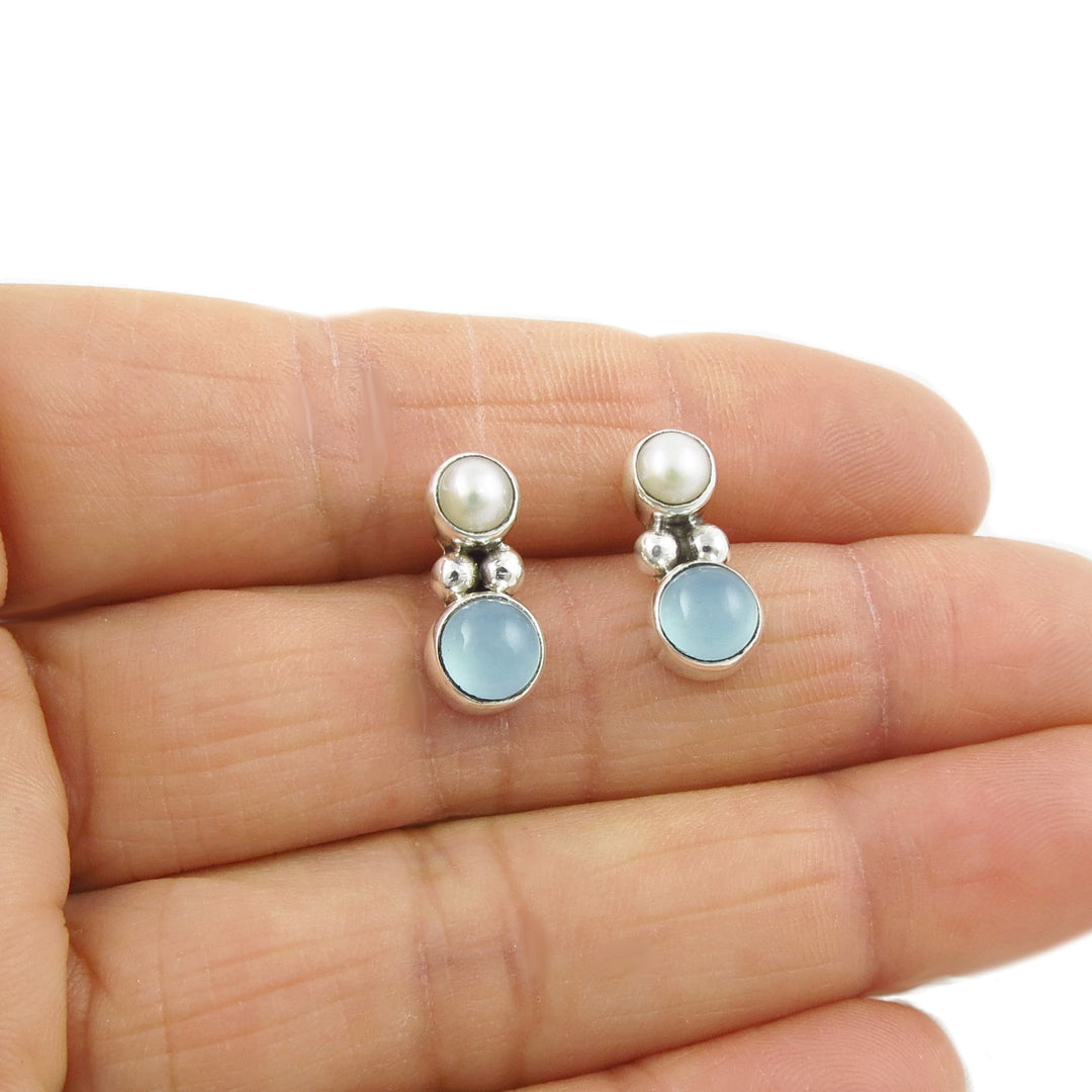 A pair of blue calcedony gemstone earrings with freshwater pearls, bezel-set in silver, held in a person's hand.