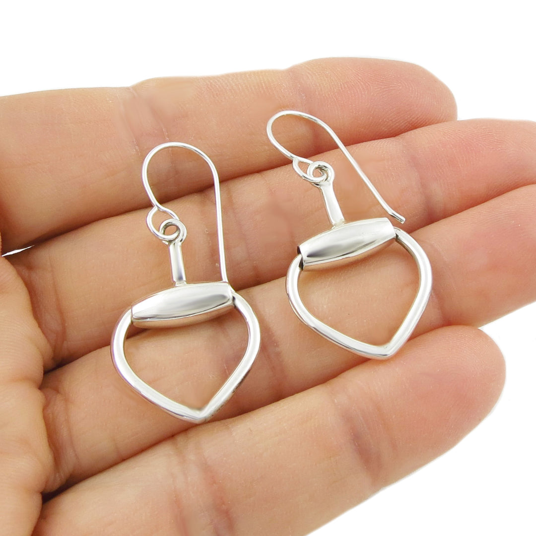 Silver heart-shaped horsebit earrings held in a hand against a white background