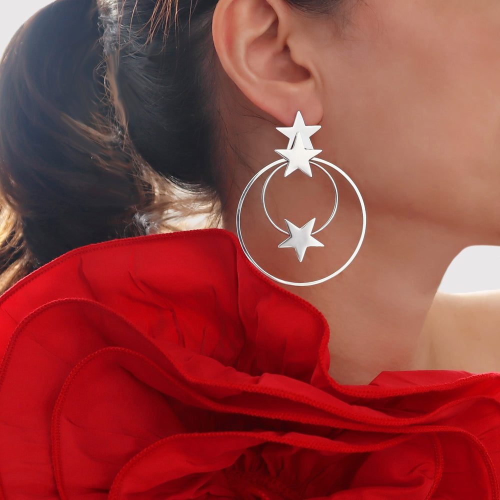Large silver hoop earrings with stars worn by a person with red fabric in the background