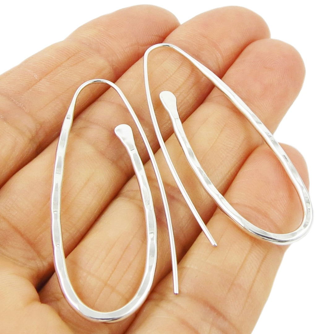 A pair of silver, handcrafted, oval drop threader earrings with a hand-hammered finish, displayed on a hand.