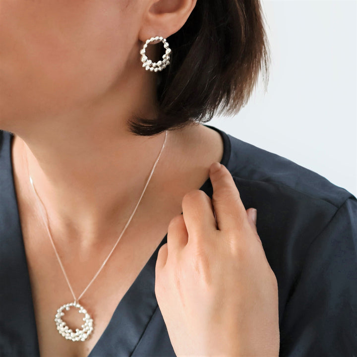 Silver earrings featuring a circle of silver bubbles with a matching neklace worn by a woman with a grey blouse on a white background