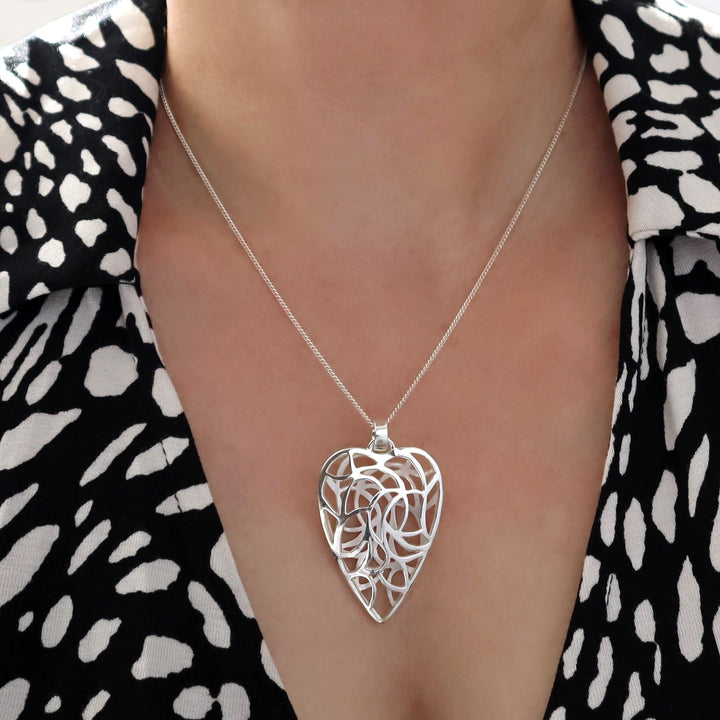 A sterling silver heart-shaped necklace with a vine pattern, worn by a person against a black and white dotted background.