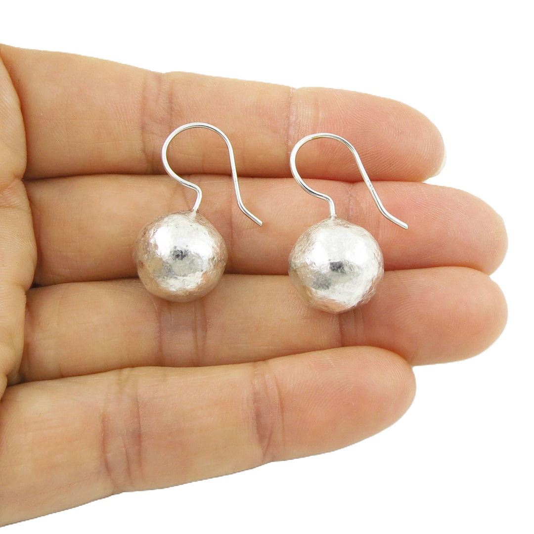 Silver ball earrings with a brutalist battered finish held between fingers on a white background