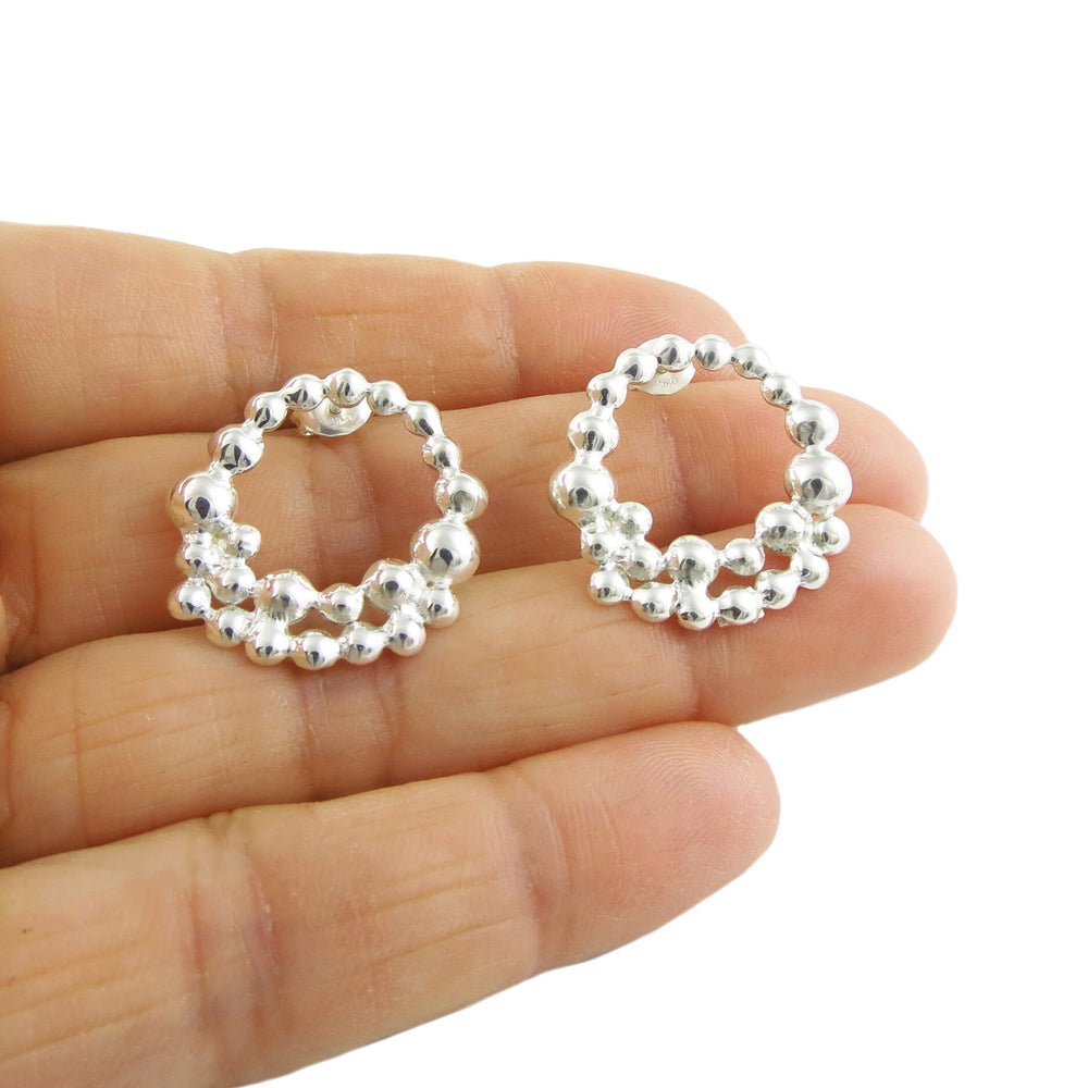 A pair of of earrings in the shape of a circle of silver bubbles being held in a persons hand on a white background