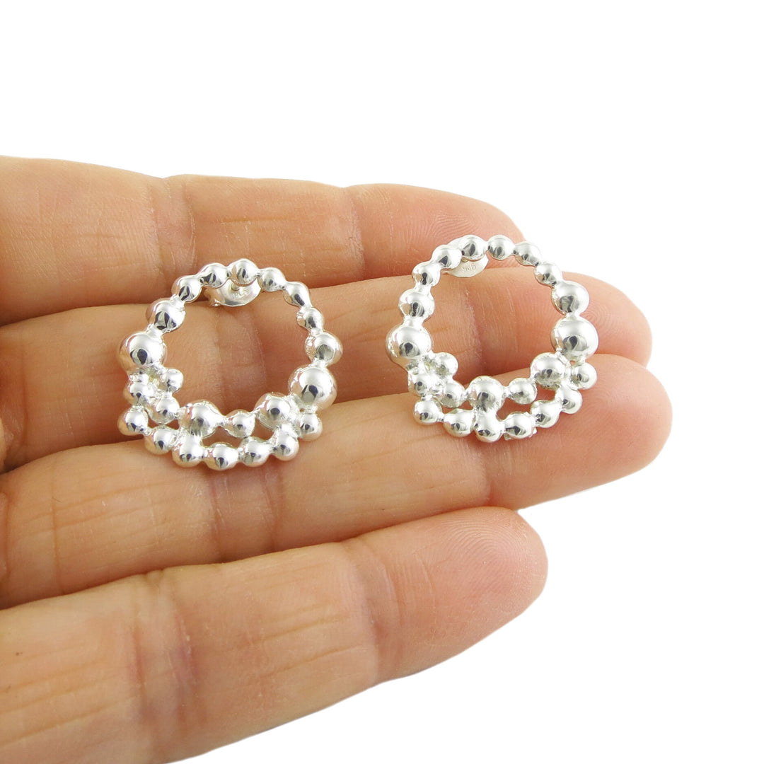 A pair of of earrings in the shape of a circle of silver bubbles being held in a persons hand on a white background