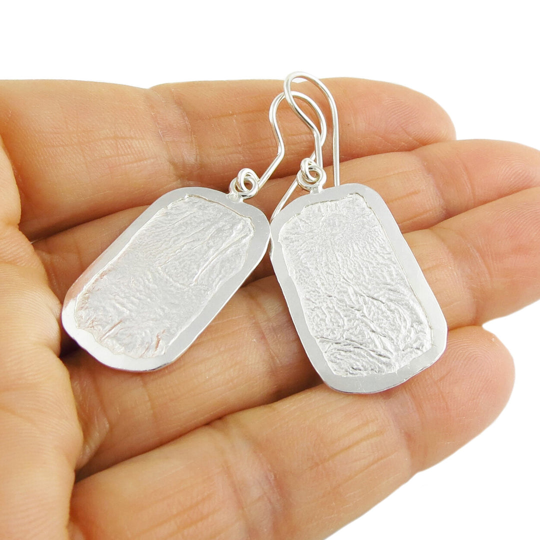 Silver earrings with a crumpled paper finish held in a person's hand.