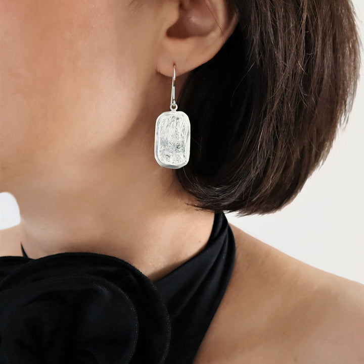 A person wearing sterling silver reticulated rectangle earrings with a matte finish and lightly polished edges.