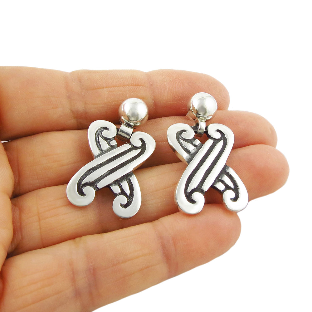 A pair of chunky Taxco silver earrings with an incised X design, held in a hand against a white background.