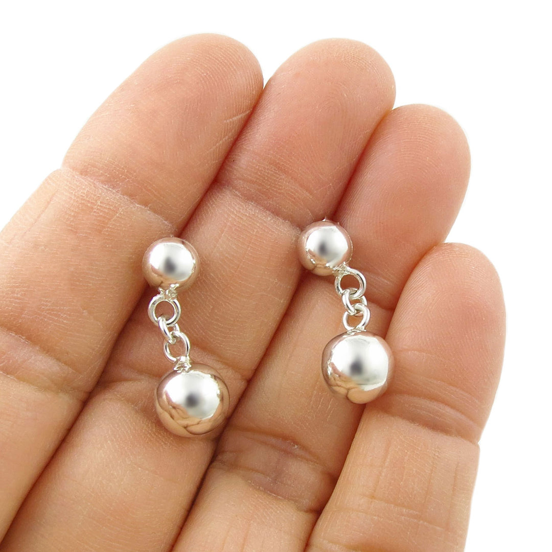 A pair of silver ball bead earrings held in a person's hand on a white background.