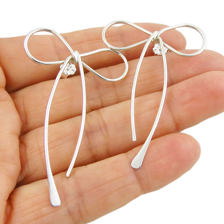 A pair of silver bow earrings with long flared ribbons, held in a person's hand.