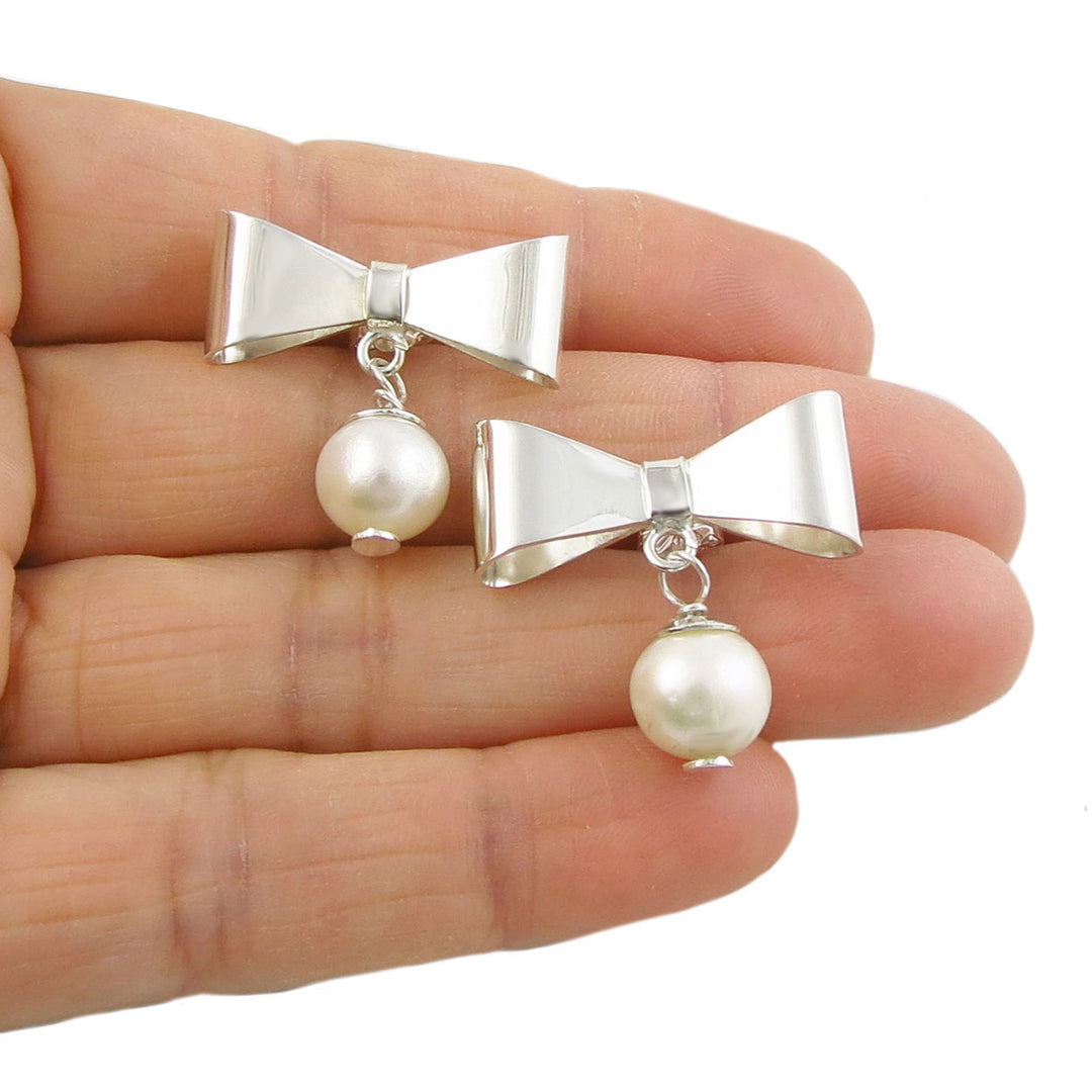 A pair of silver bow-shaped earrings with attached white pearls, held in a person's hand.