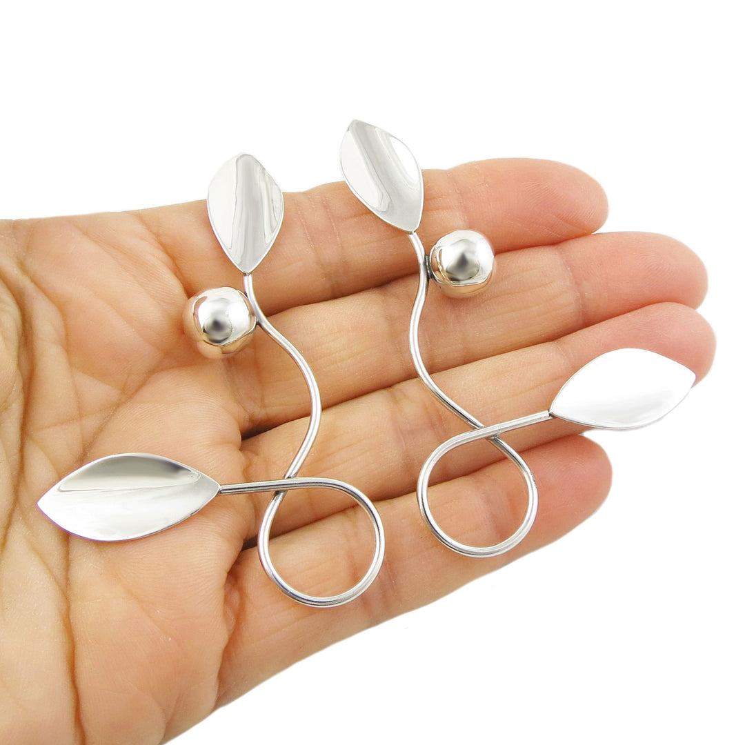 A pair of silver over-sized curling vine, leaf and berry design earrings held in a person's hand.