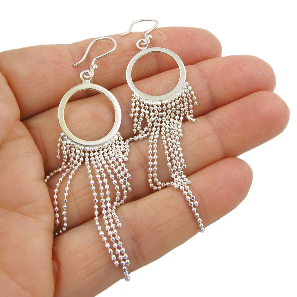 Dangling circle silver earrings with a chain fringe held in a person's hand on a white background.