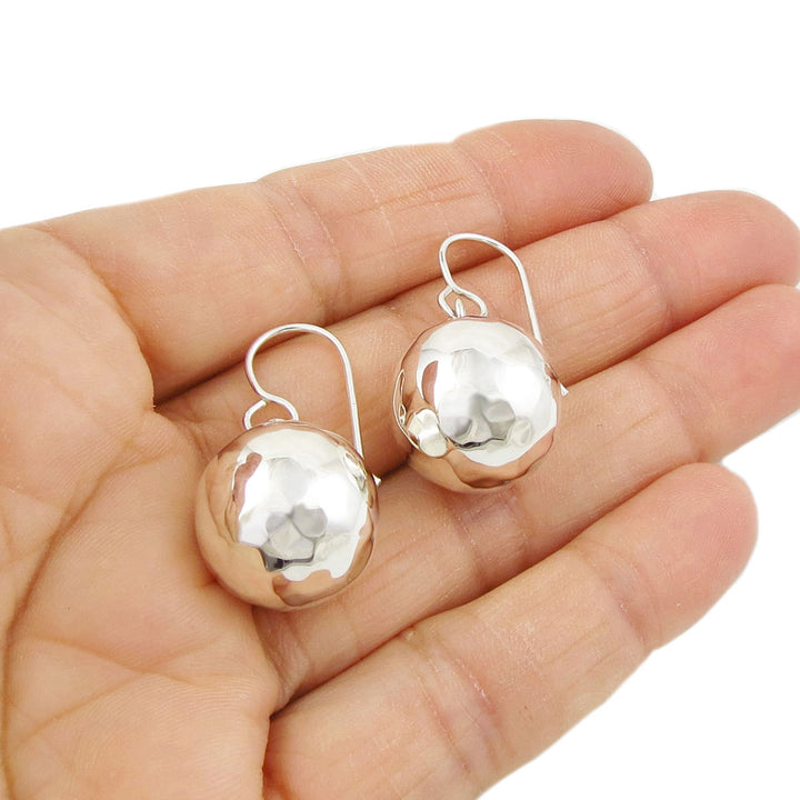 A pair of silver ball earrings with a hammered finish, held in a hand.