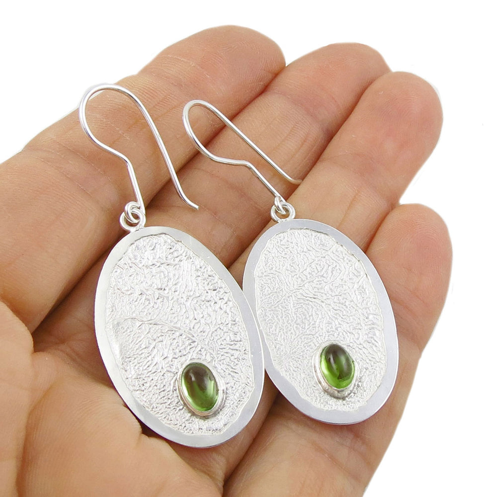 Large reticulated silvwer oval earrings set with perido gemstones held in a person's hand.