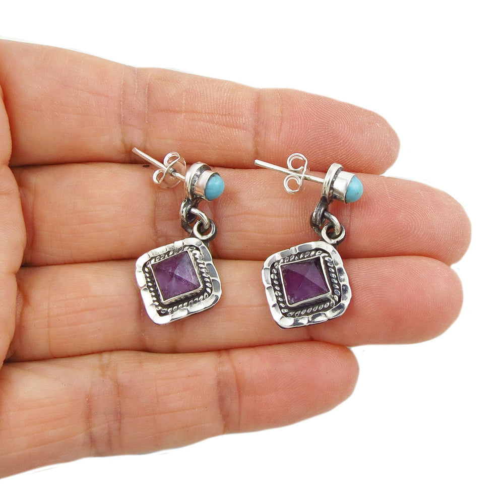 A pair of antiqued terling silver drop earrings with amethyst gemstones held in a person's hand on a white background.