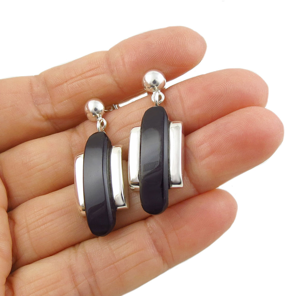 A pair of silver obsidian gemstone earrings held in a person's hand, featuring a drop design with silver accents.