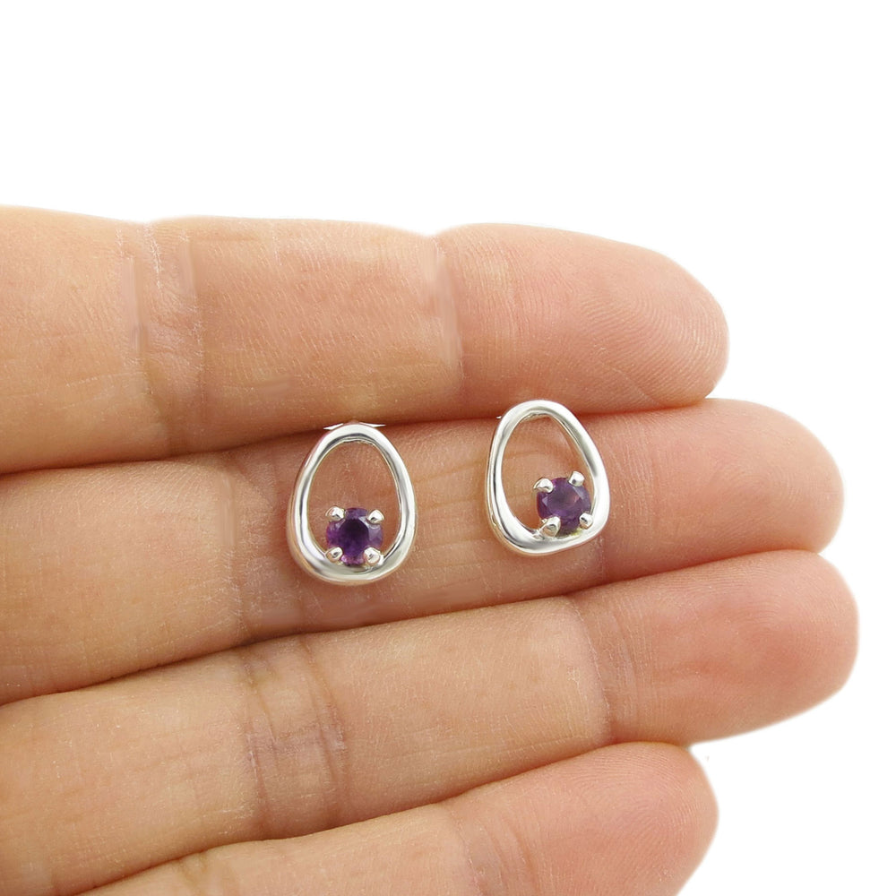 Silver earrings with purple amethyst gemstones held between fingers on a white background