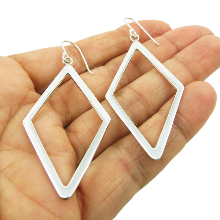 Large diamond-shaped silver earrings held in a person's hand on a white background