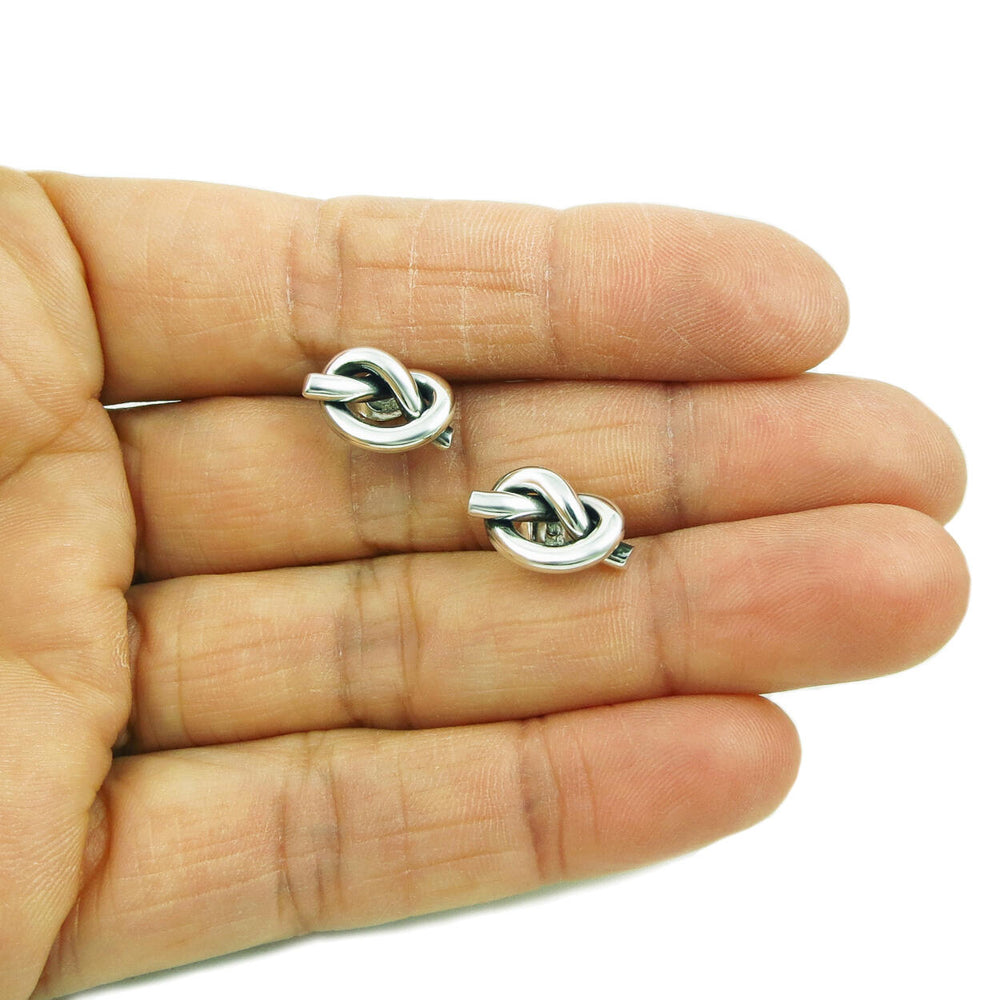 A pair of lover's knot silver stud earrings held in a person's hand on a white background