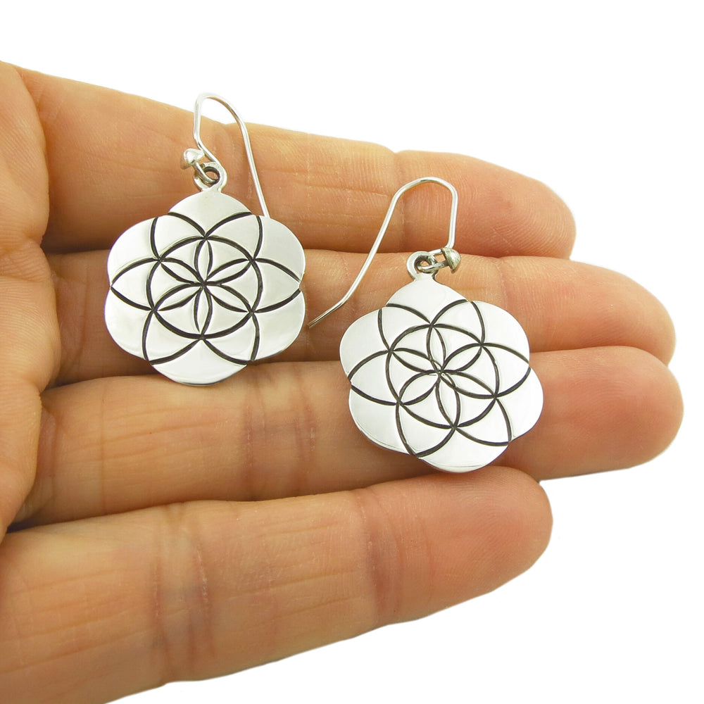 A pair of sterling silver earrings featuring the Flower of Life design, held in a person's hand.