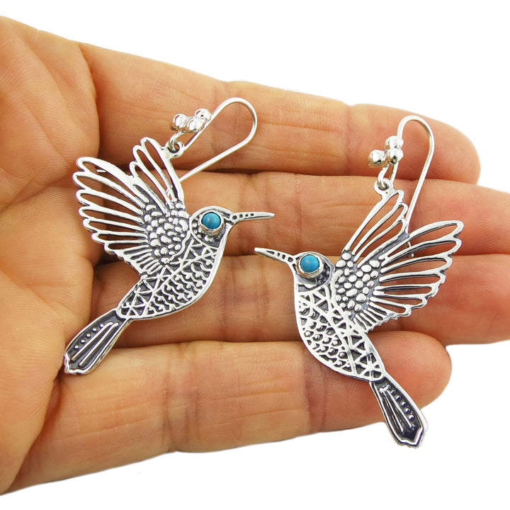 A pair of sterling silver dangle earrings in the shape of hummingbirds with turquoise-colored eyes, held in a person's hand.