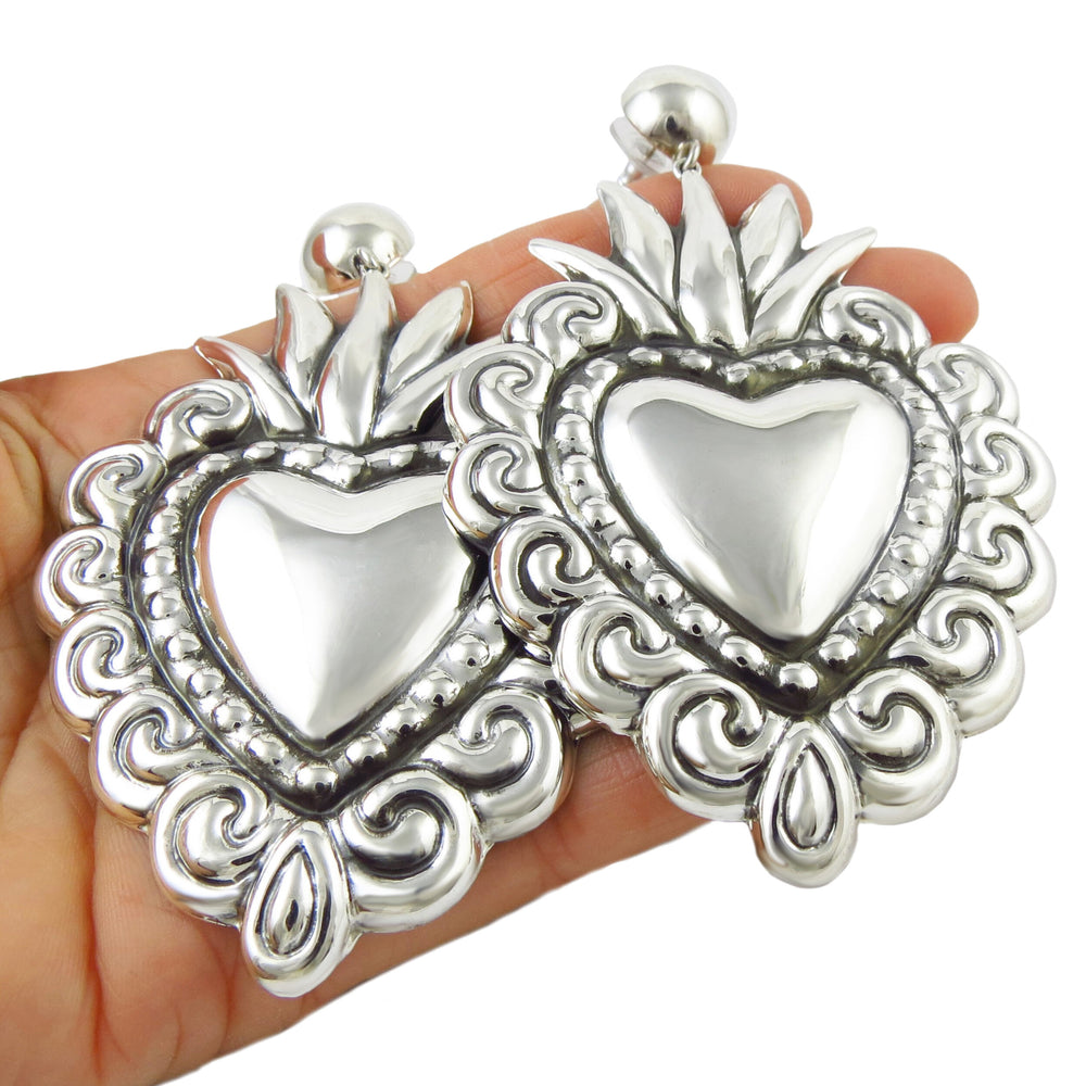 A pair of large Maria Belen Nilson designer silver earrings in the shape of heart sheld in a person's hand.