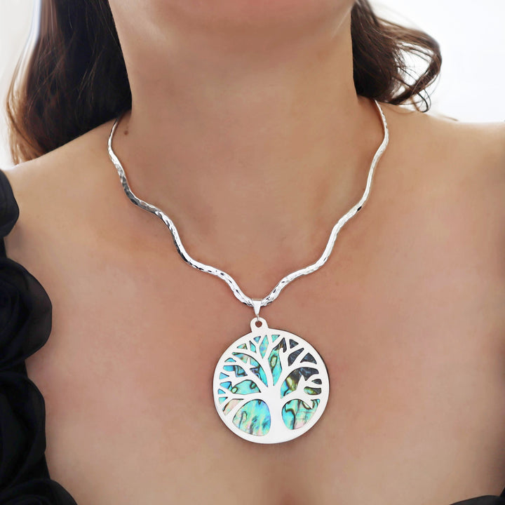 A close-up of a woman's neck and chest wearing a UK hallmarked sterling 925 silver choker necklace with a sculpted wavy design and a circular pendant featuring a tree design filled with iridescent blue-green abalone shell inlay.