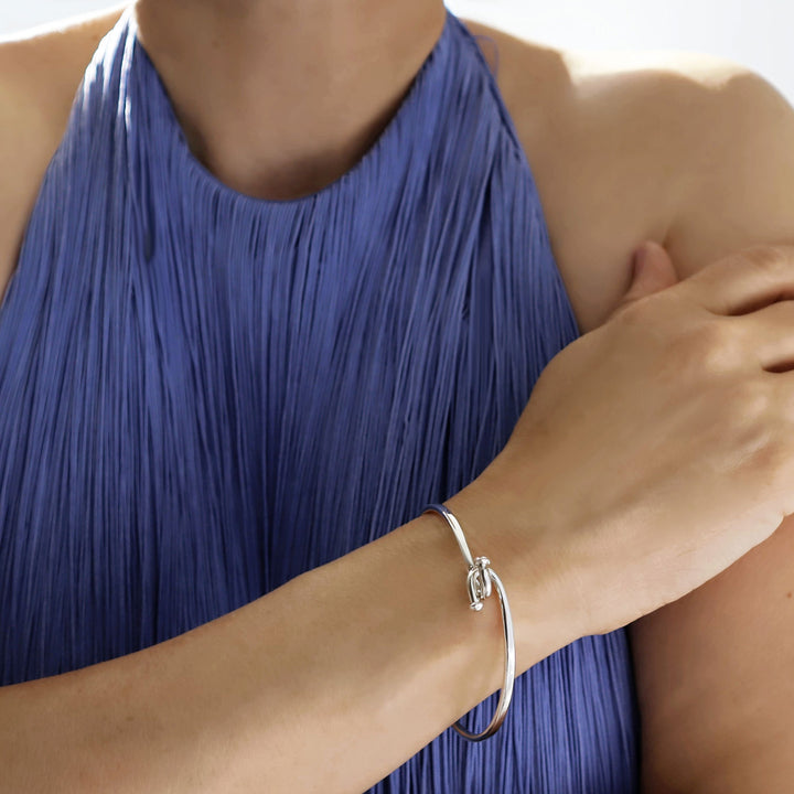 A large oval sterling silver bangle with a front twist hook feature worn by a woman in a blue dress 