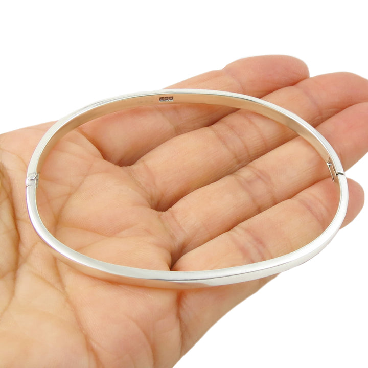 Solid silver oval bangle bracelet held in a person's hand on a white background. 