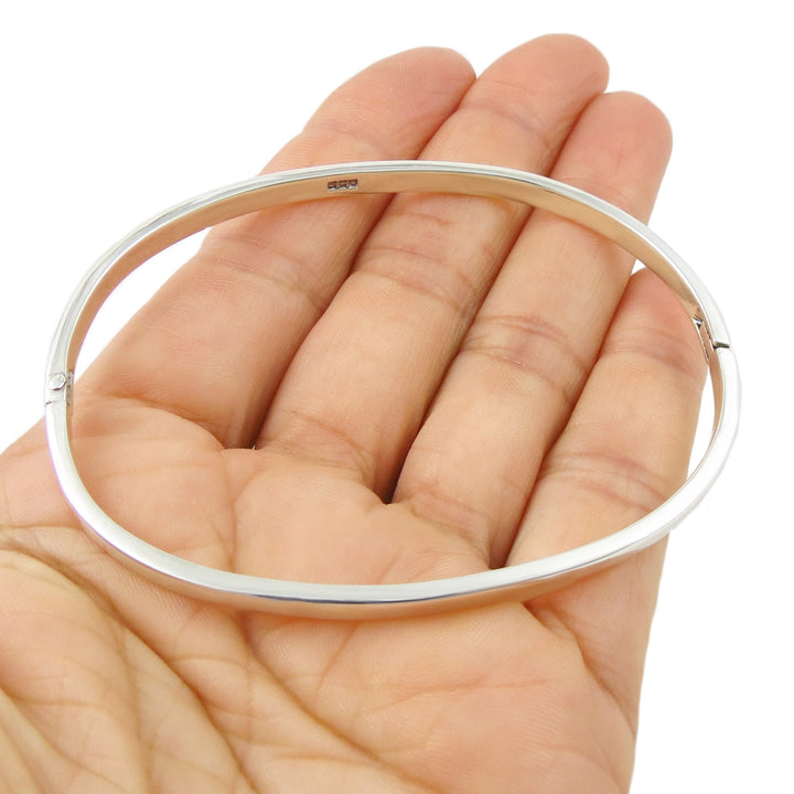 Solid silver oval bangle bracelet held in a person's hand on a white background. 
