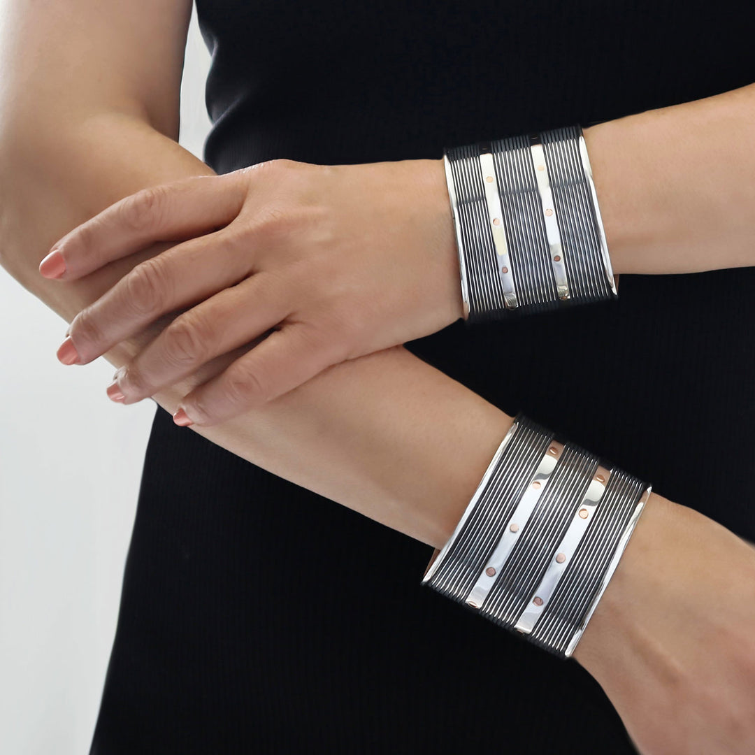 Two wide sterling silver cuff bracelets with horizontal textured bands and small two tone copper accents, shown worn on a woman's wrists against a black top and light gray background.