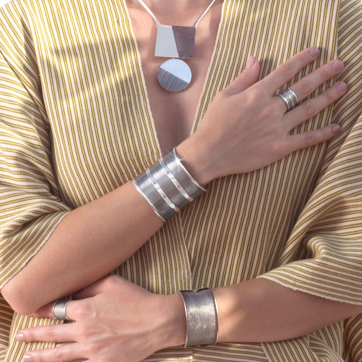 A close-up of a woman wearing two tone sterling silver and copper jewellery, including a geometric pendant necklace, multiple wide cuff bracelets, and rings, paired with a beige and brown vertically striped top.
