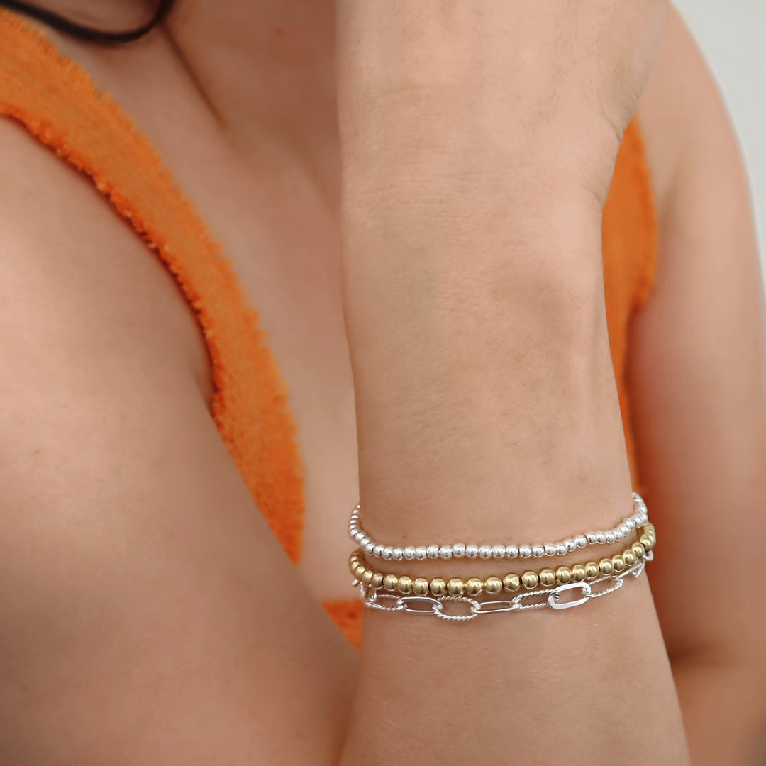 A woman in an orange top wearing a bracelet wrist stack featuring silver and gold ball bead bracelets and a paperclip chain bracelet in UK 925 sterling silver. 