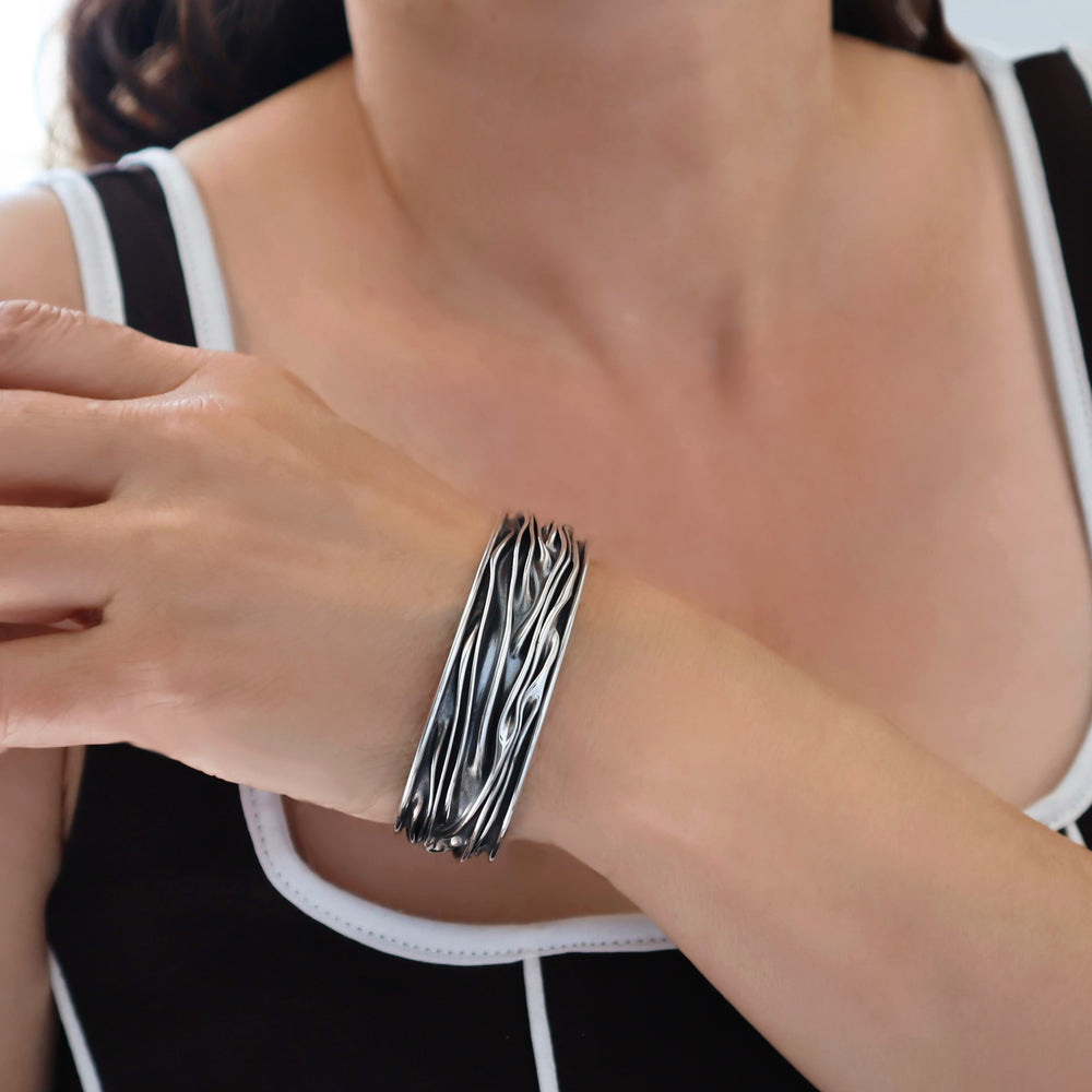 A close-up of a woman wearing a wide hallmarked crumpled sterling 925 silver cuff bracelet with a crushed design. The woman is wearing a black and white top, and the background is a clean white studio setting.
