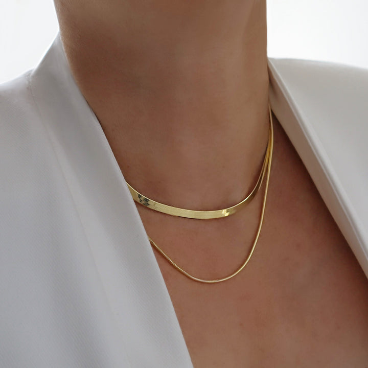 Gold snake chain and herringbone chain necklace stack worn by a person with a white jacket. 