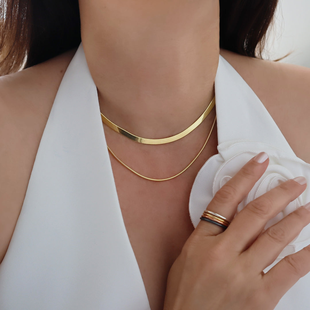 Herringbone and snake chain necklace stack in 14ct gold on a woman wearing a white garment.