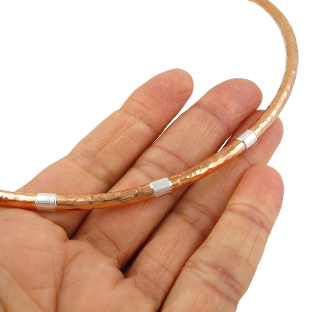 Guillermo Arregui designer hammered copper choker toruqe necklace for women with silver bands held in a person's hand on a white background. 