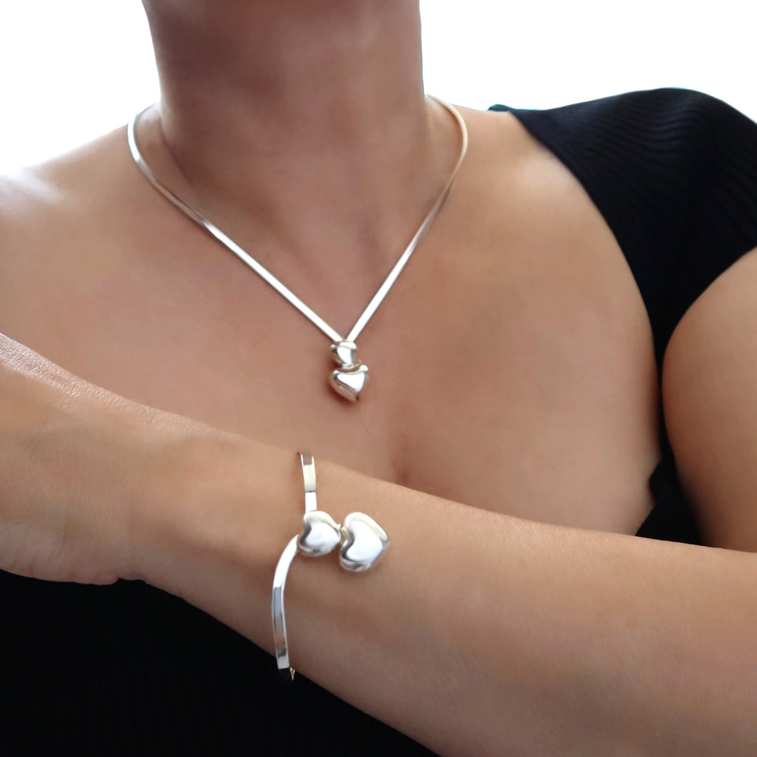 A close-up of a woman wearing a silver choker necklace with heart charms and a matching silver cuff bracelet with two heart charms, against a black top and bright white background.