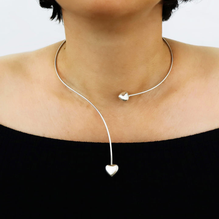 A close-up of a model's neck and upper chest wearing a delicate silver choker necklace with a curved line, a small heart at the front, and a larger dangling heart at the center of the chest.