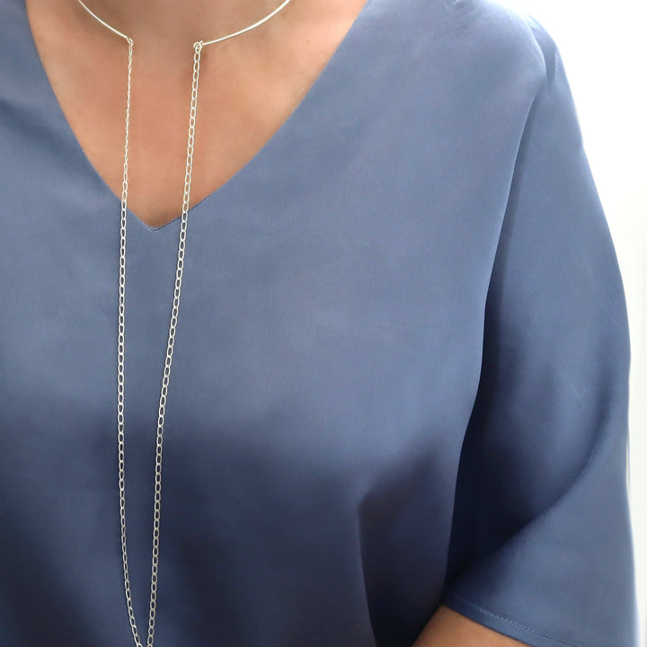 A close-up of a woman wearing a long, delicate silver chain hanging from a choker collar necklace over a slate blue V-neck top.