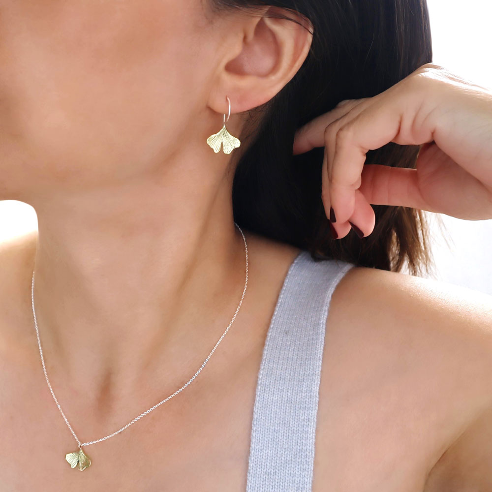 A gold and sterling silver gingko leaf earring and matching pendant and chain necklace worn by a woman with dark hair in a grey top.