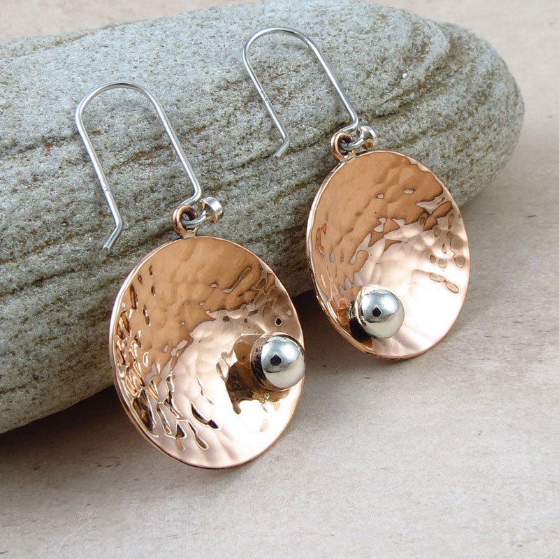A pair of two tone copper and silver earrings featuring  curved disc and beaded design with a hammered finish against a stone background