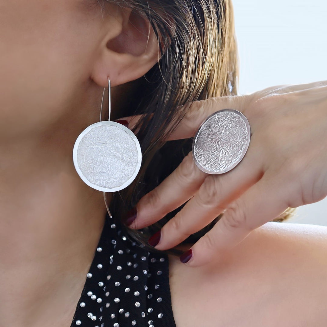 A woman wearing a large sterling silver ring and matching earring designed into large circle discs with a crumpled, rippled finish.