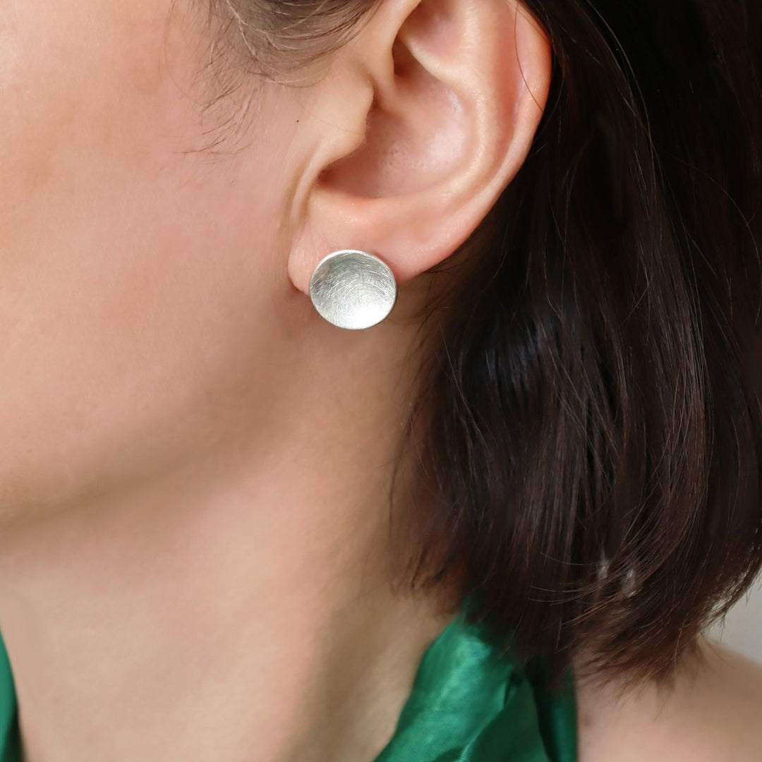 A woman with a green silk top wearing a round sterling silver stud earring with a matt, brushed finish. 