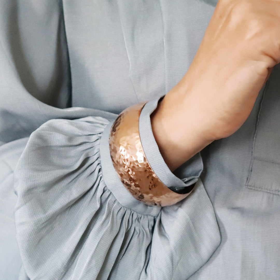 A wide hammered copper bangle worn by a woman in a blue shirt with wide sleeves
